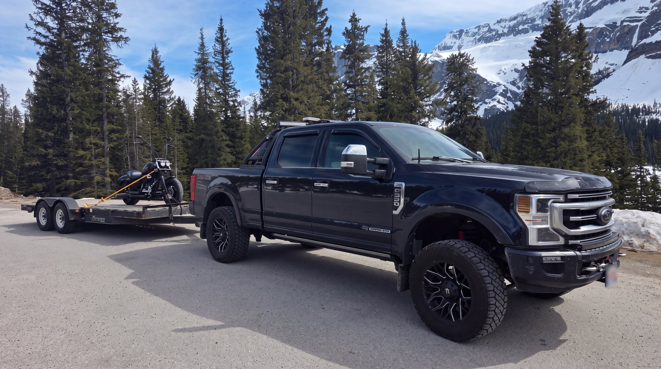 Black Ford F-350 pickup with a headache rack, light bar and beacon, towing a flat-deck trailer with snow-covered mountains behind